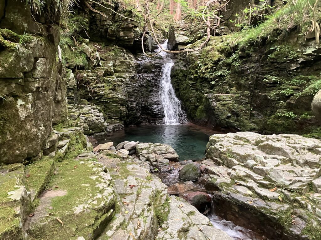緑に覆われた岩の間から流れ落ちる滝と静かなプールの風景。周囲は苔に覆われ、自然の美しさが感じられる場所。