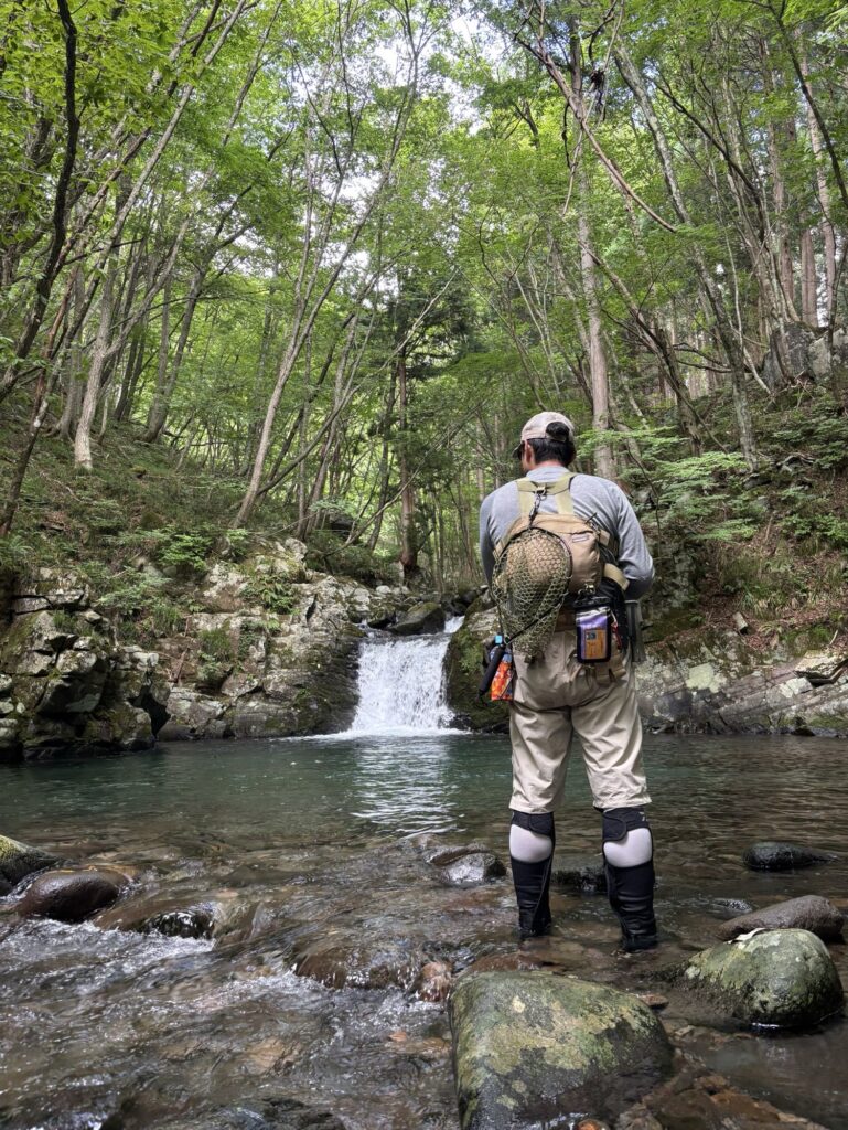 渓流で釣りをする姿が写っている。背中には渓流用のランディングネットを持ち、水辺には滑らかな岩と自然が広がっている。