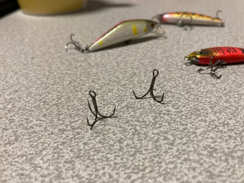 Two fishing hooks are displayed on a textured surface, with colorful lures positioned in the background.