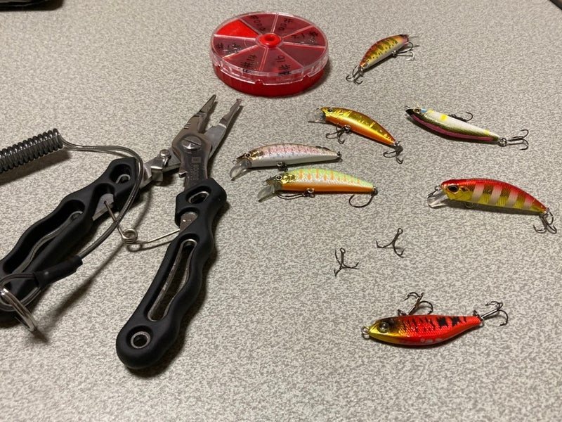 Various fishing lures arranged on a table, alongside a pair of pliers and a container of hooks.