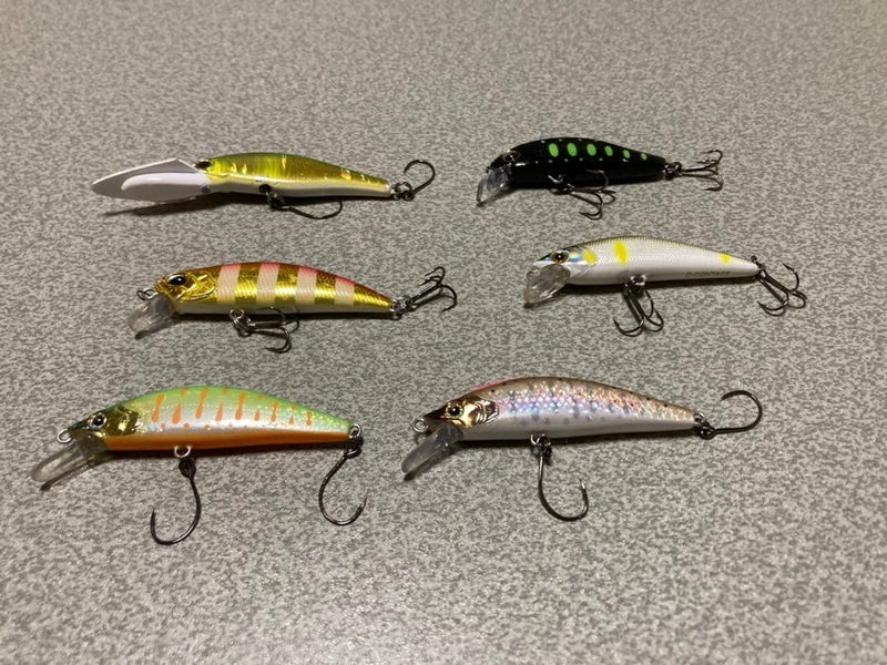 様々な色とデザインのミノーが並んでいる画像。釣り用のルアーが6つ、フック付きで、テーブルの上に置かれている。