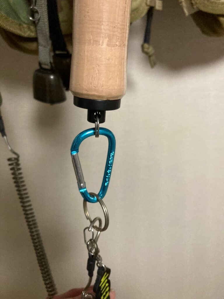 画像は、コルク製のグリップを持つフィッシングギアのハンドル部がクローズアップされています。ハンドルの底に青いカラビナと鍵リングが取り付けられており、周囲には釣り用の装備が見えます。