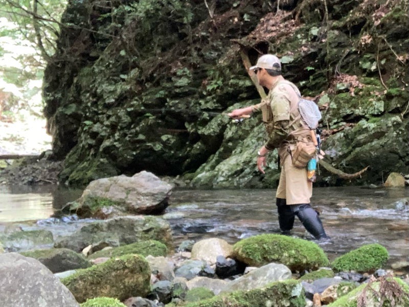 A person fishing in a stream, surrounded by rocks and greenery.