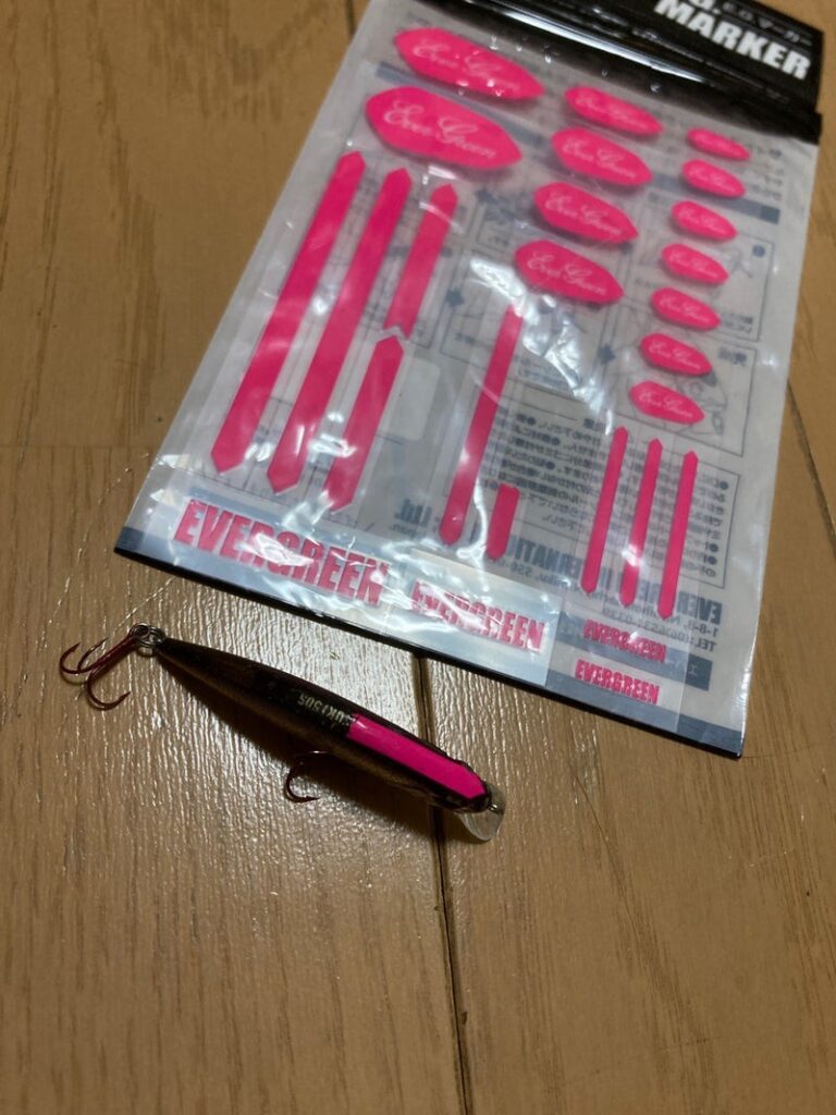 ピンク色のマーキングが施されたルアーとそのためのシールが入ったパッケージが木の床に置かれている画像