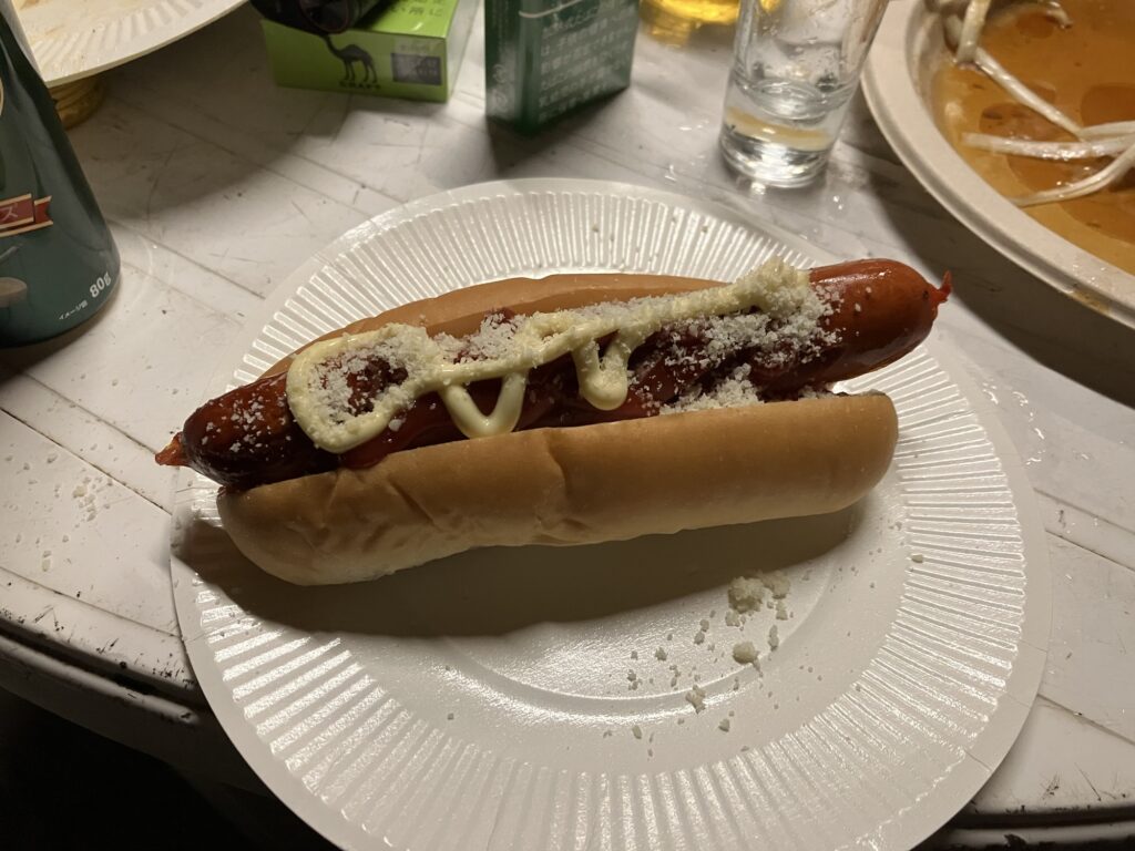 BBQで提供されたホットドッグ。ソーセージの上にマヨネーズと粉チーズがかかっている。白い紙皿に載せられている。