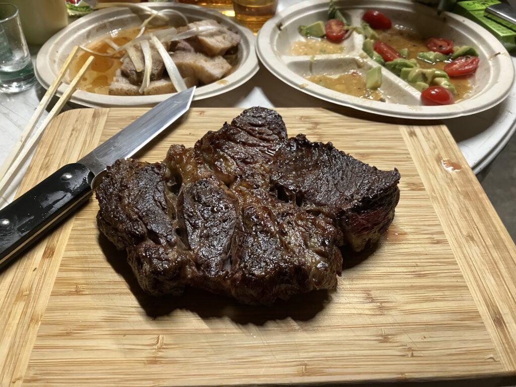 焼き加減の良いステーキが木のまな板の上に置かれ、刀がそばにあります。背景には、盛り付けられたサラダと飲み物が見えます。