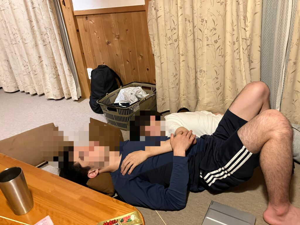 二人の男性が床に寝転がっている画像。彼らは友達のように親しげに寄り添っている。背景にはカーテンと家具が見える。