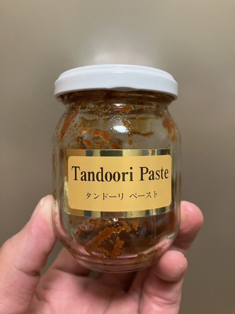 手に持たれているタンドリーペーストの瓶。瓶は透明で、ラベルには"Tandoori Paste"と"タンドーリ ペースト"の文字が書かれている。瓶の内部には使用された形跡がある。