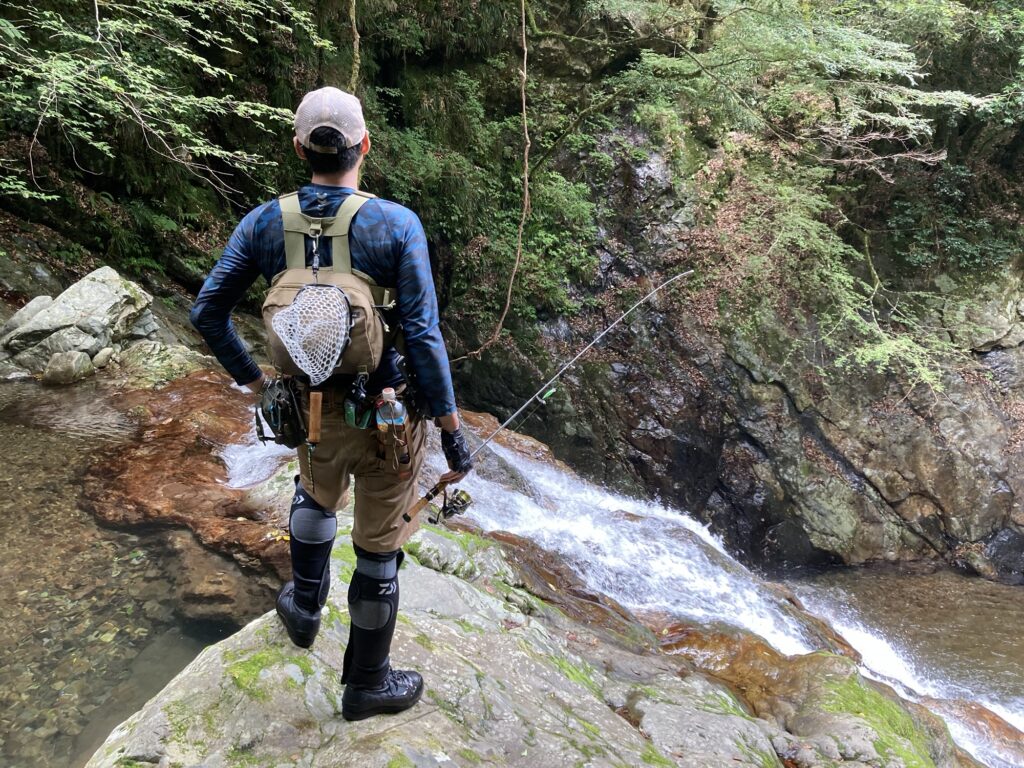 Fishing enthusiast wearing a backpack with a landing net attached, standing in a rocky creek.