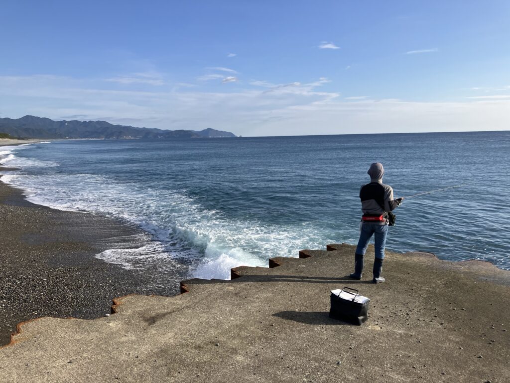 A person fishing from a small concrete ledge by the shore, with a scenic ocean view and mountains in the background. Waves gently lap at the rocky beach.