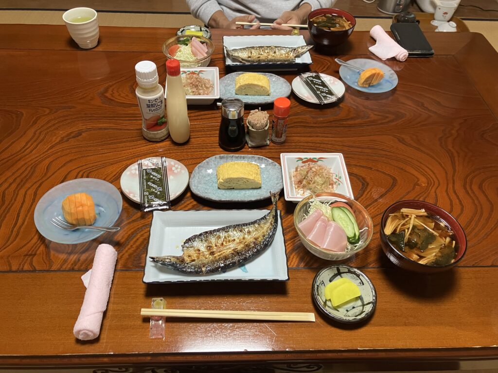和食の朝食が並べられたテーブル。焼き魚、卵焼き、刺身、味噌汁、野菜サラダなどが盛り付けられている。