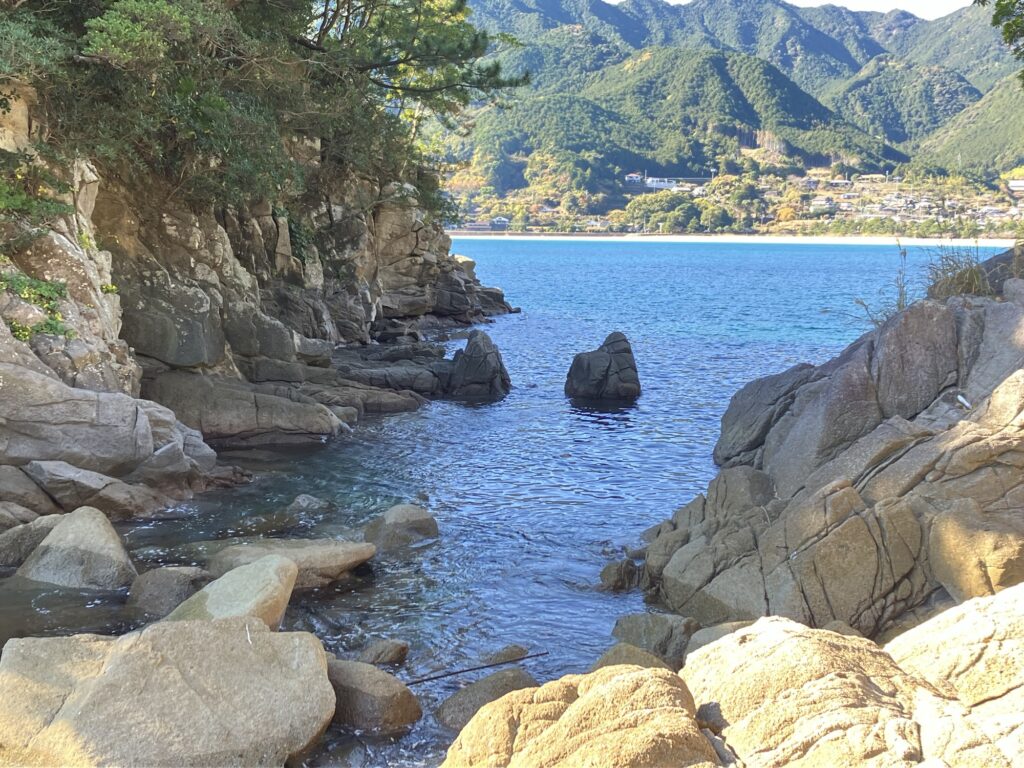 海岸の岩場と青い海が広がる風景。背後には緑豊かな山々がそびえ、静かな空間が漂う。