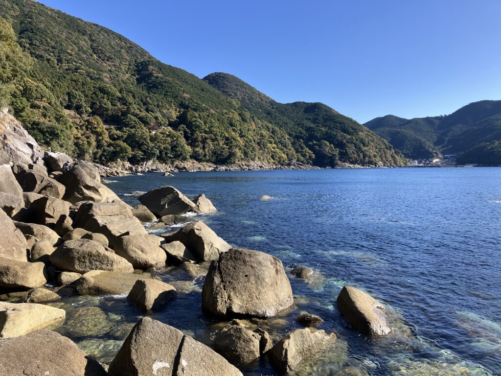 澄んだ海と岩場が広がる景色。緑の山々が背景にあり、静かな自然の風景。