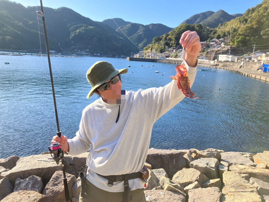 男性が釣り糸を持ち、海で釣れた赤い魚を嬉しそうに見せている。背景には山と漁港が広がり、穏やかな天候が感じられる。
